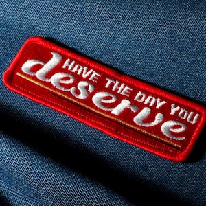 Op de afbeelding: Een rechthoekige rode geborduurde patch met de woorden "Have the day you deserve" in witte letters. De patch heeft een gouden rand en is geplaatst op een blauwe denim achtergrond.