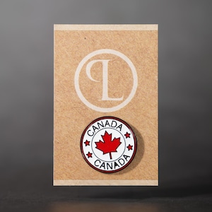 Puede incluir: Pin redondo con la palabra "CANADA" en un círculo blanco y una hoja de arce roja en el centro. El pin está colocado sobre una tarjeta beige con un logotipo "L" blanco.