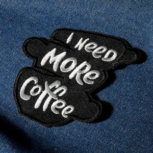Könnte beinhalten: Ein schwarzer, gestickter Aufnäher mit den Worten "I need MORE Coffee" in weißer Schrift. Der Aufnäher hat die Form von drei gestapelten Kaffeetassen und ist vor einem blauen Jeanshintergrund platziert.