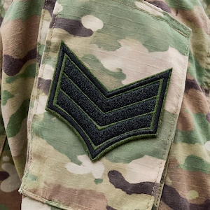 Op de afbeelding: Detail van een camouflage militair uniform met een zwart en groen chevron insigne. De patch heeft drie chevrons, die een rang aangeven. De stof van het uniform is bedrukt met een patroon van groene, bruine en beige camouflage.