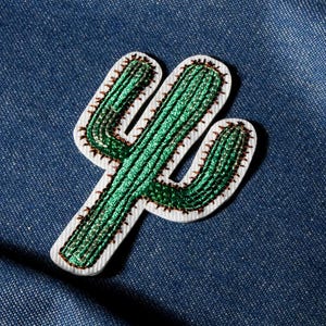 May include: A green cactus patch with white and brown trim, set against a blue denim background. The cactus design features detailed stitching and a textured appearance, perfect for adding a touch of desert flair to clothing or accessories.