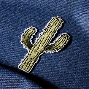 May include: Embroidered cactus patch with a white border, featuring shades of green and gold. The patch is affixed to a blue denim fabric, creating a contrast in textures and colors. This decorative patch is ideal for adding a touch of nature to clothing or accessories.