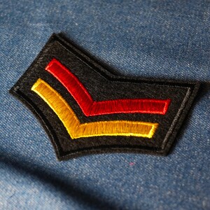 Op de afbeelding: Zwart vilten embleem met rood en geel geborduurd chevronontwerp. De patch heeft een unieke vorm en is geplaatst tegen een blauwe denim achtergrond. De patch is waarschijnlijk een embleem of insigne.