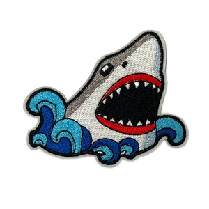 Può includere: Toppa ricamata raffigurante uno squalo con la bocca aperta, che mostra denti affilati. Lo squalo è grigio e bianco, con la bocca bordata di rosso. Onde blu circondano lo squalo, creando un tema nautico. La toppa è progettata per l'applicazione su abbigliamento o accessori.