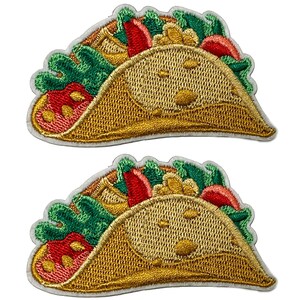 May include: Two embroidered taco patches. Each patch features a golden-yellow taco shell filled with green lettuce, red tomatoes, and other fillings. The patches have a white border.