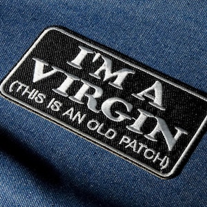 May include: Black rectangular patch with white embroidered text reading "I'M A VIRGIN (THIS IS AN OLD PATCH)". The patch has a white border and is set against a blue denim background. A humorous accessory.
