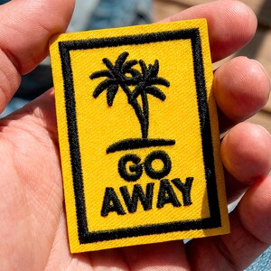Può includere: Toppa rettangolare gialla con bordo nero, con un disegno di palma nera e le parole "GO AWAY" in nero. La toppa è tenuta in una mano.