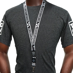 Könnte beinhalten: Ein dunkelgraues T-Shirt mit einem Sicherheitsbeauftragten-Lanyard. Auf dem Lanyard steht "SECURITY OFFICER". Auf beiden Ärmeln des T-Shirts sind weiße Logos zu sehen.