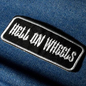 Może przedstawiać: Czarna prostokątna naszywka z białym haftowanym napisem "HELL ON WHEELS". Naszywka ma białą obwódkę i jest umieszczona na niebieskim tle z dżinsu. Tekst jest w gotyckiej czcionce.