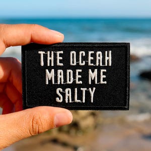May include: A black rectangular patch with white embroidered text reading "THE OCEAN MADE ME SALTY." The patch is held up against a blurred background of the sea and rocks.