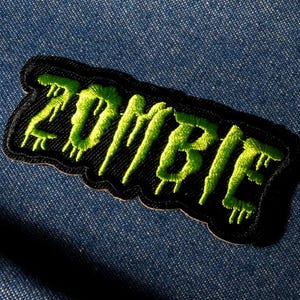 Peut inclure: Un écusson noir brodé avec le mot "ZOMBIE" en lettres vert fluo dégoulinantes. L'écusson a une bordure noire et est posé sur un fond en denim bleu. Un accessoire sur le thème d'Halloween.