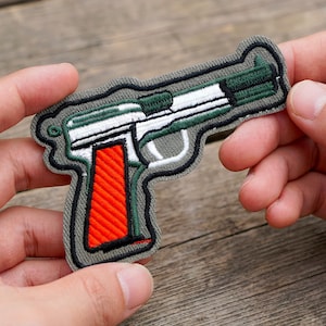 Op de afbeelding: Geborduurde patch in de vorm van een handpistool. De patch heeft een grijze omtrek, een witte slede, een groene loop en een feloranje handgreep. Het ontwerp is gedetailleerd met zwarte stiksels.