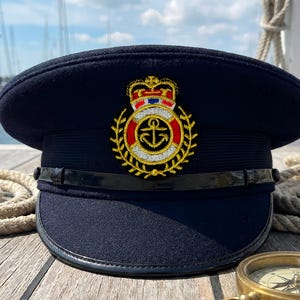 May include: A navy blue naval officer's hat with a gold and red emblem featuring an anchor, crown, and laurel wreath. The hat has a black visor and band. The background includes a wooden surface, rope, and a compass.