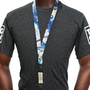 Peut inclure: Un cordon avec un motif de vagues bleues et blanches, porté autour du cou d'une personne. Le cordon a un fermoir noir et une petite étiquette avec du texte japonais. La personne porte un t-shirt gris foncé avec un logo blanc sur les manches.