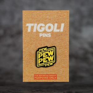 Può includere: Un prodotto Tigoli Pins su una scheda marrone. La spilla è nera e gialla con il testo "PEW PEW PEW" in un carattere stilizzato. La scheda ha anche il testo "TIGOLI PINS" e "FOR ENTERTAINMENT PURPOSES ONLY".