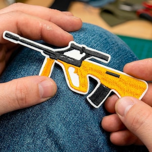 May include: Embroidered patch of a yellow and black rifle with a scope. The patch has a white border and is held against a blue denim background. The rifle design is detailed with black and white accents.