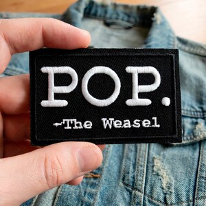 Könnte beinhalten: Schwarzer rechteckiger Aufnäher mit weiß gesticktem Text „POP.“ und darunter „-The Weasel“. Der Aufnäher wird vor einer Jeansjacke gehalten.