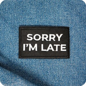 Può includere: Una toppa rettangolare nera con la scritta bianca "SORRY I'M LATE". La toppa è posta su uno sfondo di denim blu, creando un contrasto di trame e colori. La toppa è probabilmente un accessorio da stirare o cucire.