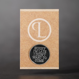 Op de afbeelding: Een zwarte en zilveren ronde pin met de tekst "SORRY I'M LATE I DIDN'T WANT TO COME" wordt weergegeven op een bruine kaart met een wit "P" logo. De pin is een humoristisch accessoire.