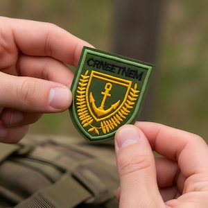 May include: A green and gold embroidered patch with the text "CRNEETNEM" at the top. The patch features a gold anchor in the center, surrounded by a gold wreath. The patch is held between two hands.