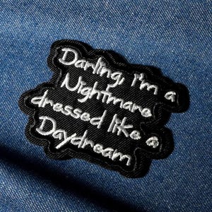 Könnte beinhalten: Ein schwarzer, gestickter Aufnäher mit weißem Schreibschrift-Text, der lautet: "Darling I'm a Nightmare dressed like a Daydream." Der Aufnäher hat einen schwarzen Rand und befindet sich auf einem blauen Jeans-Hintergrund.