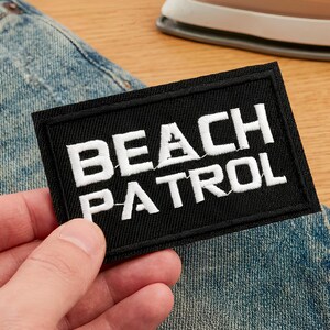 Könnte beinhalten: Schwarzer rechteckiger Aufnäher mit weiß gesticktem Text „BEACH PATROL“. Der Aufnäher befindet sich über einer blauen Jeans. Ein Bügeleisen ist im Hintergrund zu sehen.