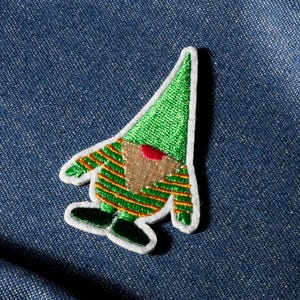 Può includere: Toppa ricamata a forma di gnomo con cappello verde, camicia a righe verdi e arancioni, barba marrone e scarpe nere. La toppa ha un bordo bianco su sfondo denim blu.