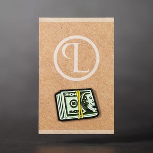 Pode incluir: Um alfinete com uma pilha de notas de 100 dólares americanos presa com uma faixa amarela. O alfinete está sobre um fundo bege com um desenho circular branco e a letra "L".