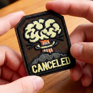 May include: A brown embroidered patch with a cartoon explosion design and the word "CANCELED" in yellow. The patch has a black border and is held by two hands.