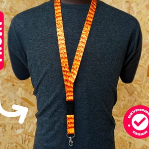 Pode incluir: Um cordão com um padrão repetitivo em forma de chama em tons de laranja e amarelo. O cordão é usado ao redor do pescoço e tem um fecho de plástico preto e um clipe de metal. A palavra "FRONT" está impressa em um rótulo vermelho.