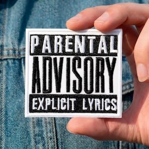 Può includere: Una toppa quadrata bianca con testo nero che recita "PARENTAL ADVISORY EXPLICIT LYRICS". La toppa è tenuta contro una giacca di jeans. Il testo è in grassetto, senza grazie.