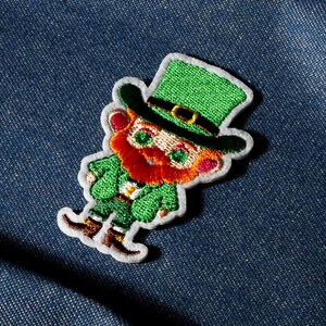 Pode incluir: Patch bordado com um duende de terno e chapéu verdes com barba ruiva. O patch tem uma borda branca e está sobre um fundo de ganga azul. O duende é um tema do Dia de São Patrício.