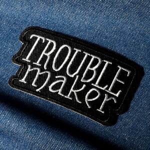 Pode incluir: Um patch bordado em preto e branco com as palavras "TROUBLE MAKER" em escrita branca. O patch é retangular com cantos arredondados e está sobre um fundo de ganga azul.