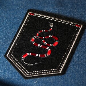 Peut inclure: Un écusson noir brodé d'un motif de serpent rouge et blanc. L'écusson est bordé de coutures blanches et d'un accent rouge. Il est posé sur un fond en denim bleu.