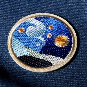 Peut inclure: Écusson brodé représentant une nuit étoilée, avec des fils bleus, blancs et dorés. Le patch rond a une bordure beige et est posé sur un fond en denim bleu. Le motif représente un ciel nocturne tourbillonnant.
