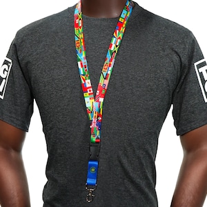 Könnte beinhalten: Ein Schlüsselband mit einem Muster aus internationalen Flaggen hängt um den Hals einer Person. Das Schlüsselband hat einen schwarzen Verschluss und einen blauen Akzent. Die Person trägt ein dunkelgraues T-Shirt mit einem weißen Logo auf dem Ärmel.