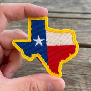 Op de afbeelding: Geborduurde patch in de vorm van de staat Texas, met het ontwerp van de vlag van Texas. De patch heeft een blauw veld met een witte ster en een witte, rode en gele rand. De patch wordt tussen twee vingers gehouden.