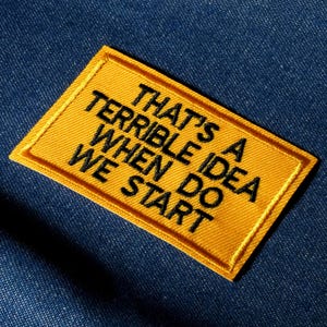 May include: A yellow rectangular patch with black embroidered text reading "THAT'S A TERRIBLE IDEA WHEN DO WE START" is set against a blue denim backdrop. The patch has a yellow border and is slightly angled.