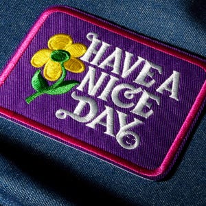 May include: Embroidered patch with a purple background, a pink border, and the words "HAVE A NICE DAY" in white script. A yellow flower with green leaves is in the upper left corner. The patch is rectangular with rounded corners.