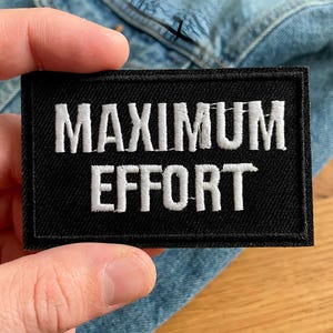 May include: A black rectangular patch with the words "MAXIMUM EFFORT" embroidered in white. The patch is held in a hand, with a denim jacket in the background. The patch has a black border.