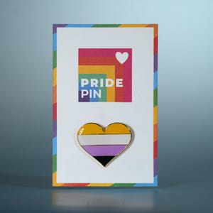 Op de afbeelding: Een hartvormige non-binaire pride pin met goud, wit, paars en zwart email. De pin wordt weergegeven op een kaart met een regenboogrand en de woorden "PRIDE PIN". Bovenop staat een regenboogvierkant met een wit hart.