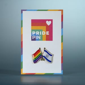 Può includere: Una spilla Pride su una carta bianca con un bordo arcobaleno. La carta presenta le parole "PRIDE PIN" e un simbolo a forma di cuore. Sono attaccate due spille, una con una bandiera arcobaleno e l'altra con la bandiera israeliana.