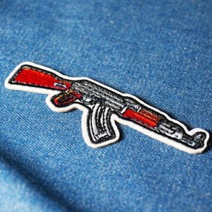 Könnte beinhalten: Gestickter Aufnäher mit einer detaillierten Darstellung eines AK-47 Sturmgewehrs. Der Aufnäher hat einen weißen Rand, wobei der Körper des Gewehrs in Grau- und Schwarztönen gehalten ist und rote Akzente aufweist. Der Aufnäher befindet sich auf einem blauen Jeans-Hintergrund.