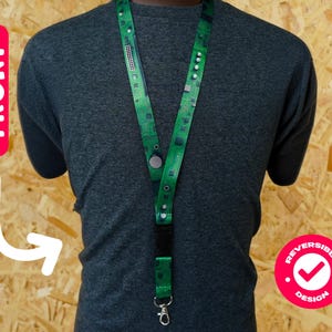 Può includere: Lanyard verde con un design a circuito stampato, indossato intorno al collo. Ha una chiusura nera, una clip in metallo e una chiusura a bottone. La parola "FRONT" è su un'etichetta rossa. Il design è reversibile.