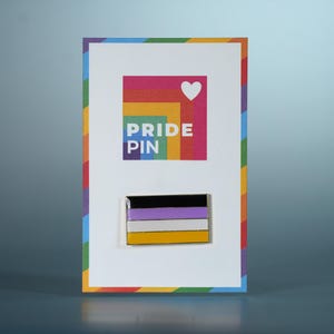 May include: A non-binary pride pin on a card with a rainbow border. The pin features a black, white, purple, and yellow flag design. The card has the words "PRIDE PIN" and a heart symbol.