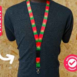 Puede incluir: Un cordón rojo, verde y blanco con el diseño de la bandera portuguesa. Tiene un cierre de plástico negro y un clip metálico. El diseño es reversible. El cordón se muestra en una persona que lleva una camiseta gris.