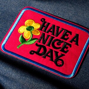 Peut inclure: Écusson brodé avec un fond rouge, une bordure bleue et les mots "HAVE A NICE DAY" en noir. Une fleur jaune avec des feuilles vertes est sur le côté gauche de l'écusson. L'écusson est rectangulaire avec des coins arrondis.