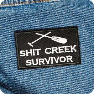 May include: A black rectangular patch with white text and a crossed oar and paddle design. The text reads "SHIT CREEK SURVIVOR". The patch is sewn onto a blue denim fabric.