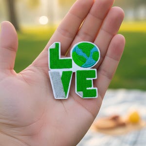 May include: Embroidered patch spelling "LOVE" with a green and white color scheme. The "O" is a globe with blue oceans and green continents. The patch is held in a hand against a blurred outdoor background.