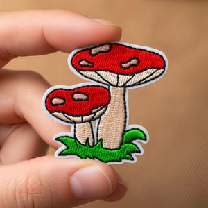 May include: Embroidered patch featuring two red and white mushrooms with beige stems and green grass. The patch is held between fingers against a tan background. The mushrooms have a detailed design.
